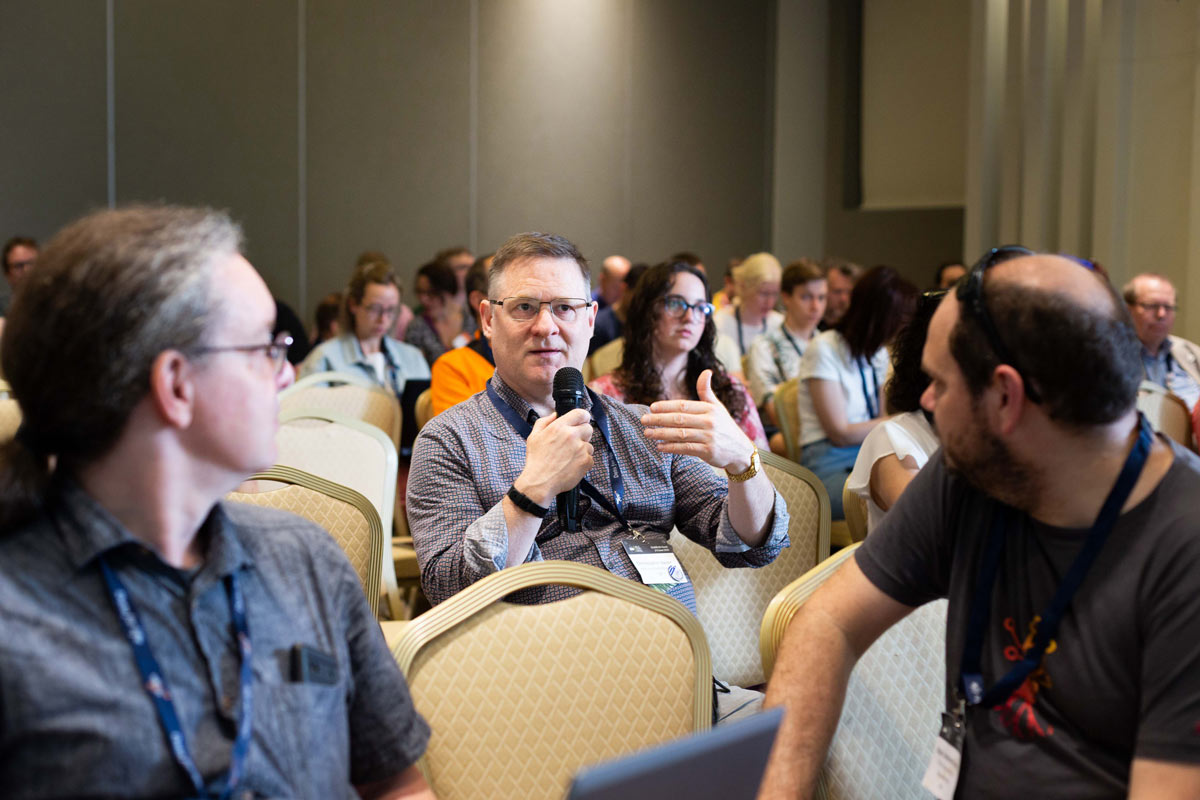 Participants of Elixir All Hands 2025 discuss at a workshop, a man holds a microphone and talks, while two other man look at him.